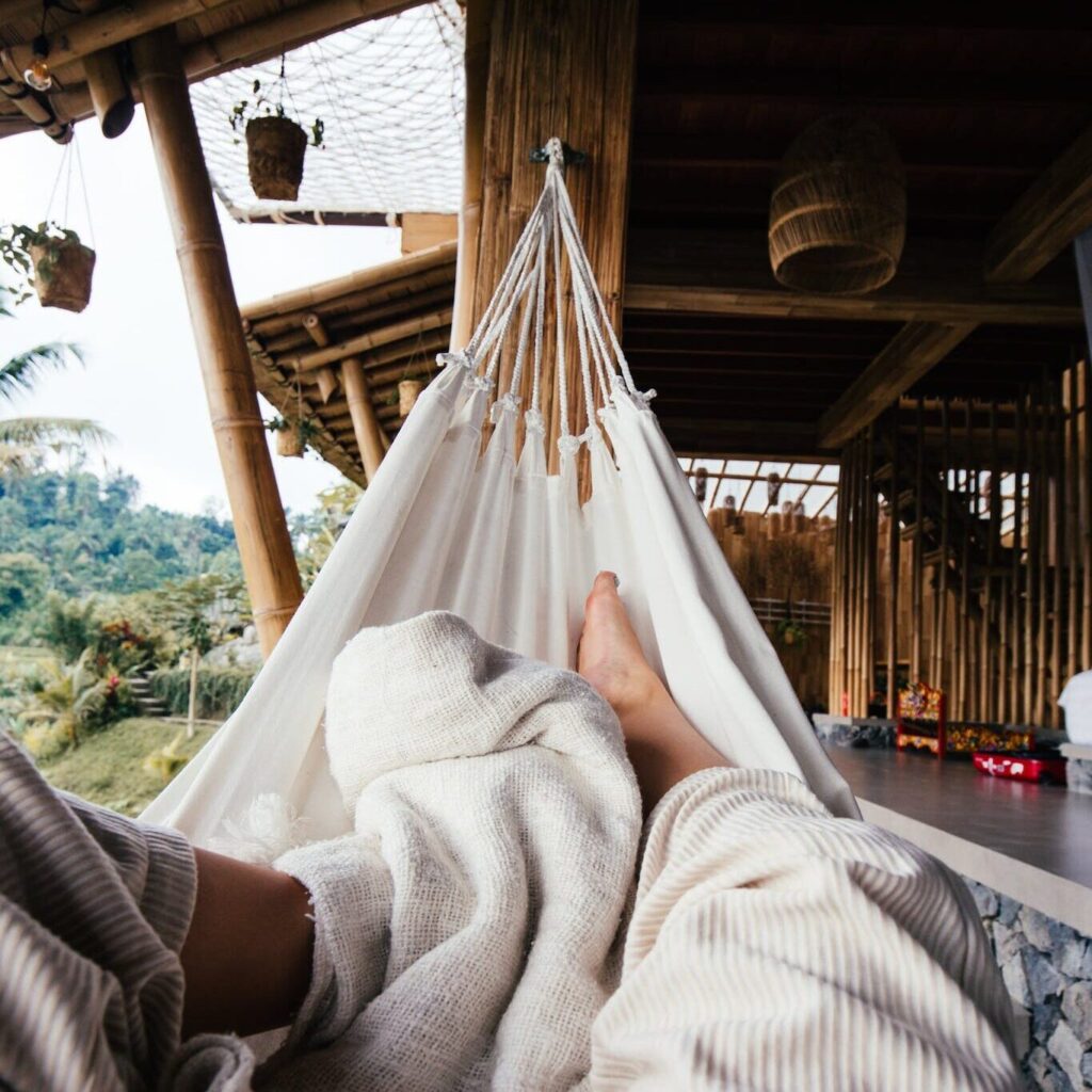 Crop barefoot traveler chilling on white cozy hammock under blanket on terrace of house in daytime