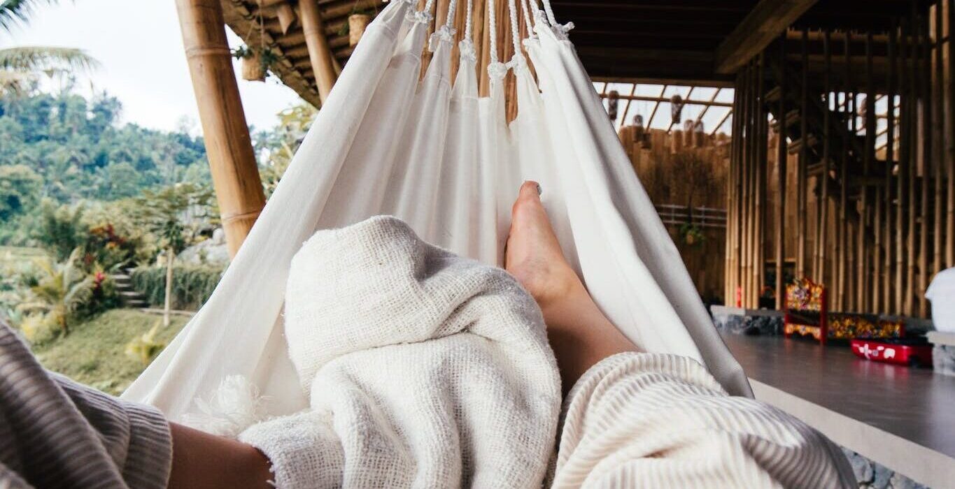 Crop barefoot traveler chilling on white cozy hammock under blanket on terrace of house in daytime